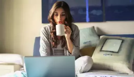 A woman on her laptop while sitting in bed, sipping on her morning coffee and enjoying her association's microlearning programs